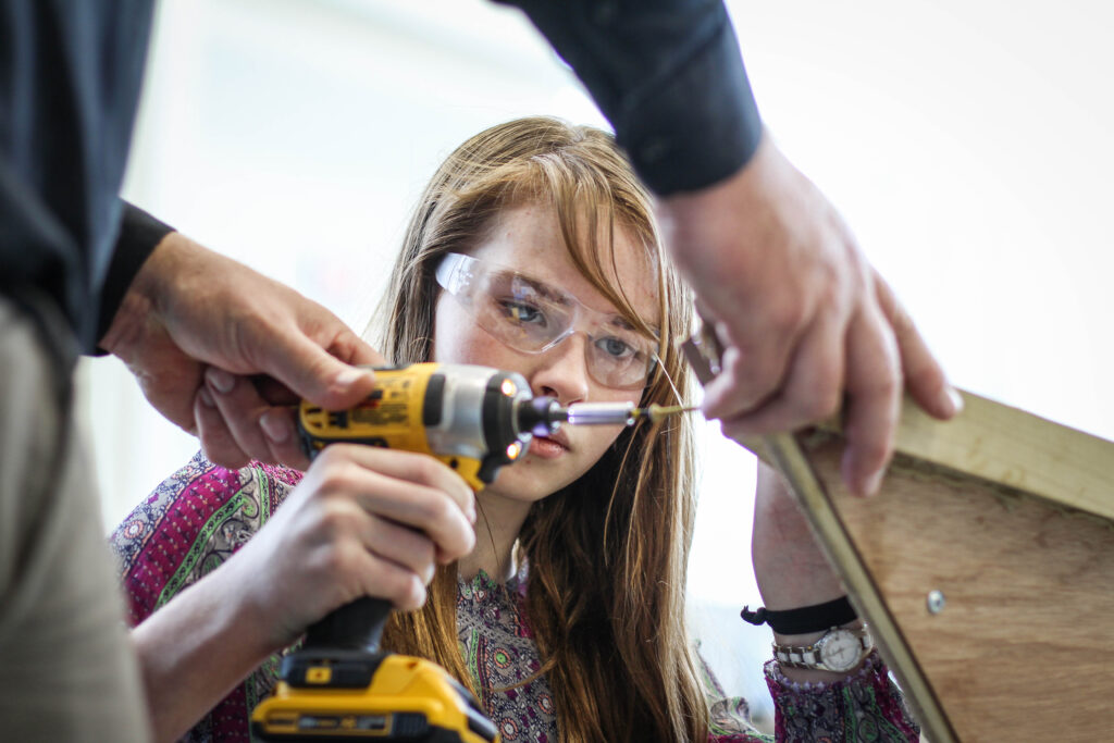 Person helping a girl use a drill on a wooden board.