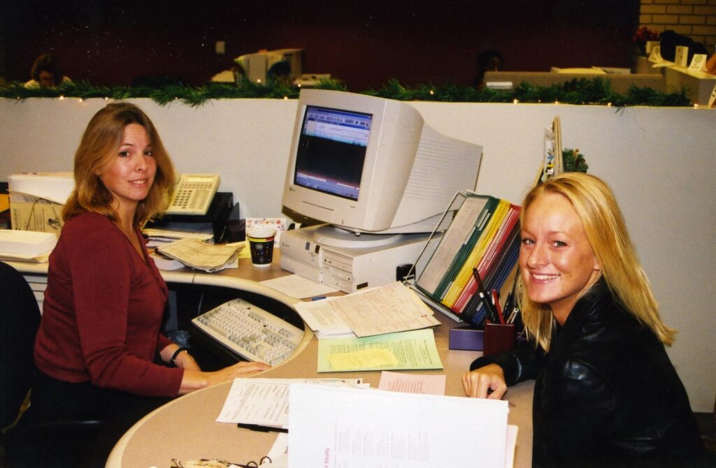 Two women working near a computer while smiling at the camera.