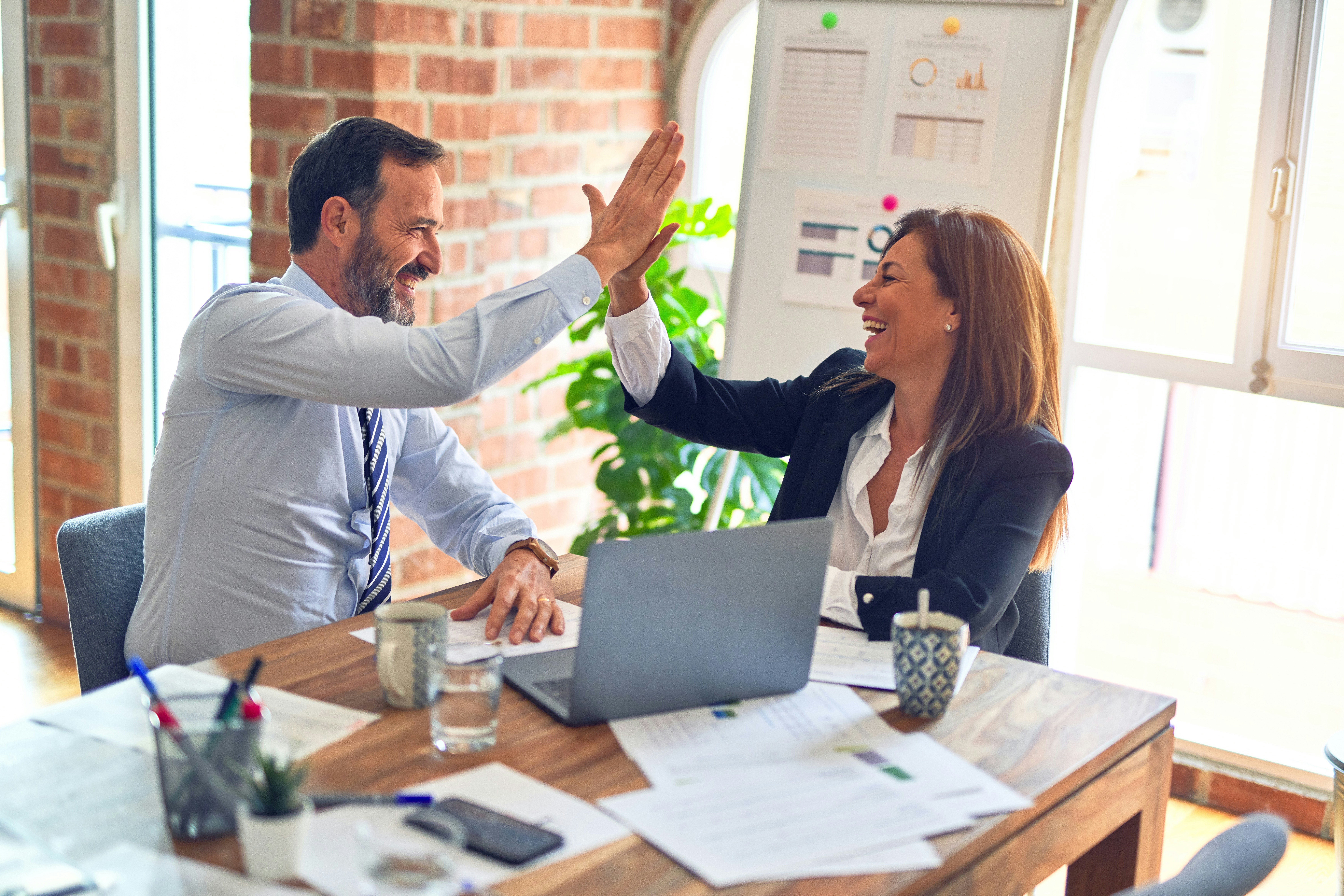 Two business people high-fiving over a table with papers and laptop