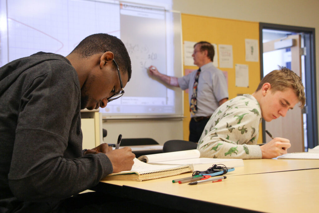 Two students working at a desk with a teacher doing math on a whiteboard behind them